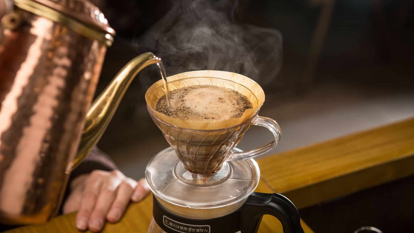 A hand pours hot water from a copper kettle into a coffee dripper filled with ground coffee. The dripper is placed on top of a mug. Steam rises from the brewing coffee, and a wooden countertop is visible in the background.