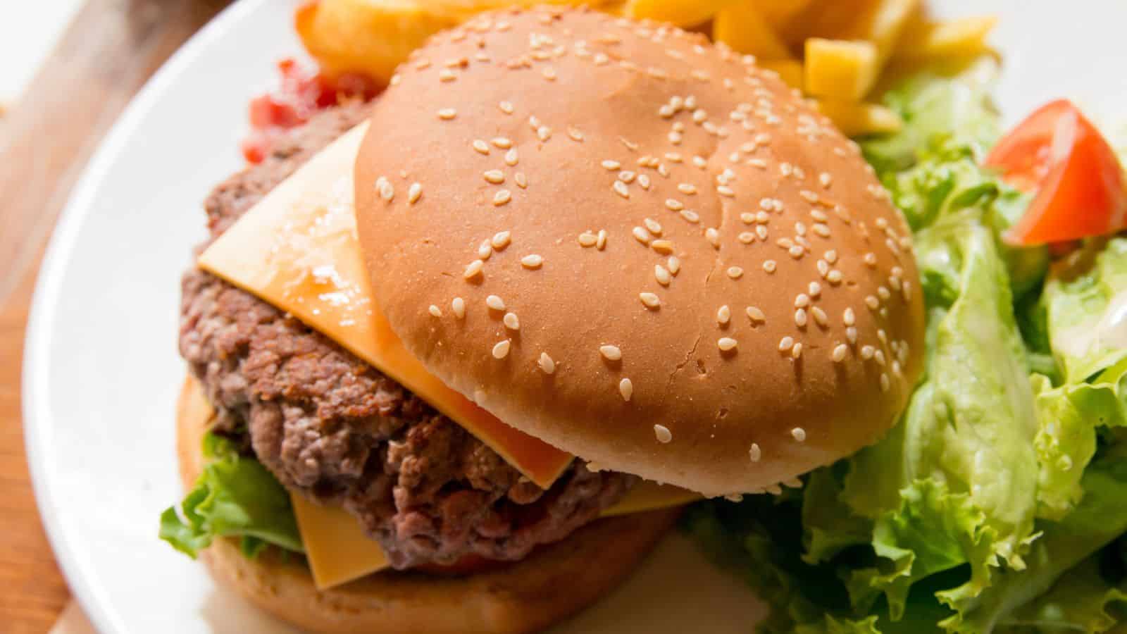 A hamburger with a sesame seed bun, a beef patty, and cheese is served on a white plate. It is accompanied by lettuce, a wedge of tomato, and French fries in the background.