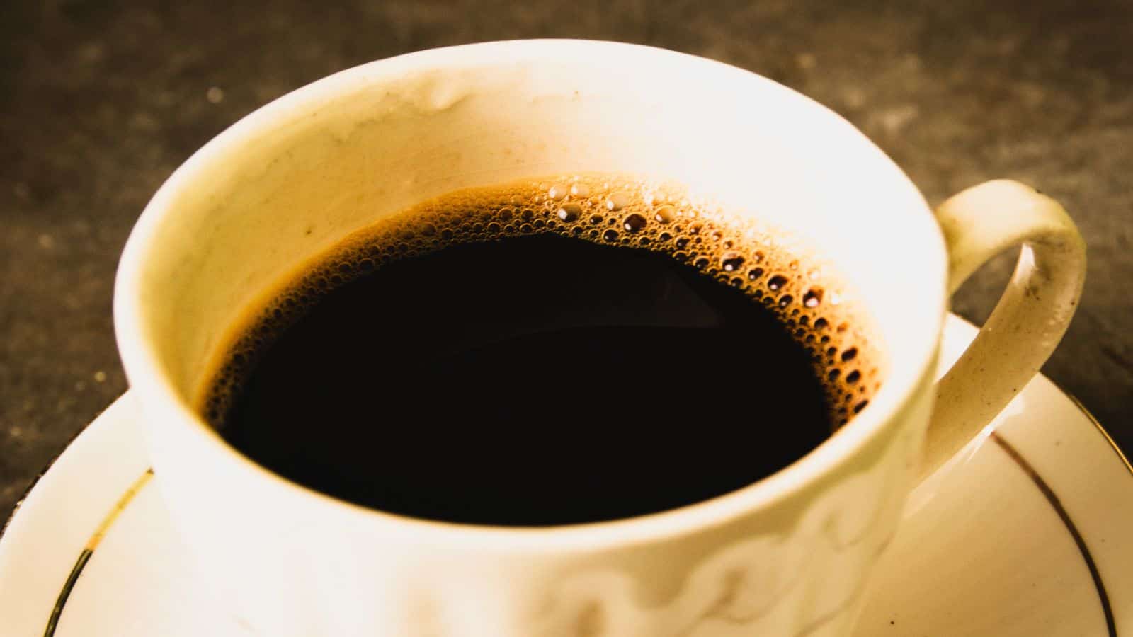 A close-up image of a white cup filled with black coffee. The cup is on a matching saucer, and the coffee has a layer of small bubbles on its surface. The background is a dark, textured surface.