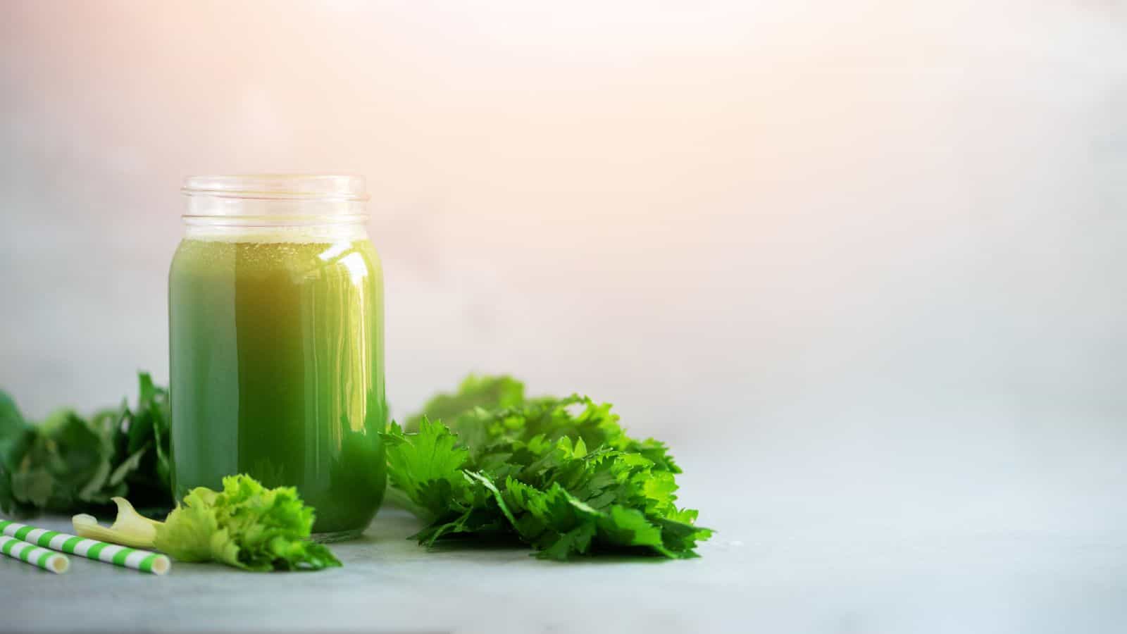 A mason jar filled with green juice is placed on a light surface. Fresh cilantro leaves are scattered around the jar. Two striped green and white paper straws are next to the jar. The background is softly blurred.