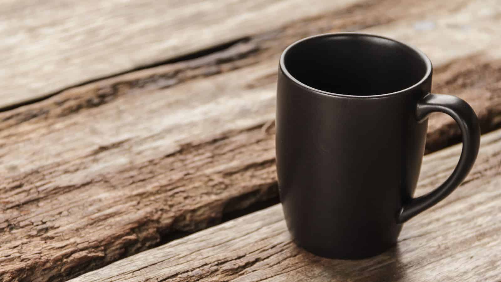 A black ceramic mug is placed on a rustic wooden surface. The mug is empty, and its handle is on the right side. The wood's texture is rough and weathered, creating a contrasting background for the smooth surface of the mug.
