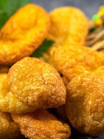A close-up of a pile of golden-brown fried snacks placed on a gray surface. A few green leaves garnish the top. Colorful skewers are visible in the background.