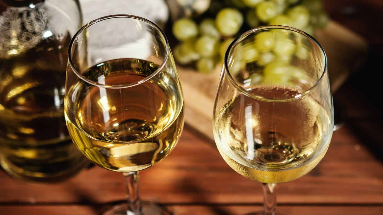 Two glasses of white wine are placed on a wooden table. A wine bottle is partially visible in the background, alongside a bunch of green grapes. The setting suggests a relaxed, informal atmosphere.