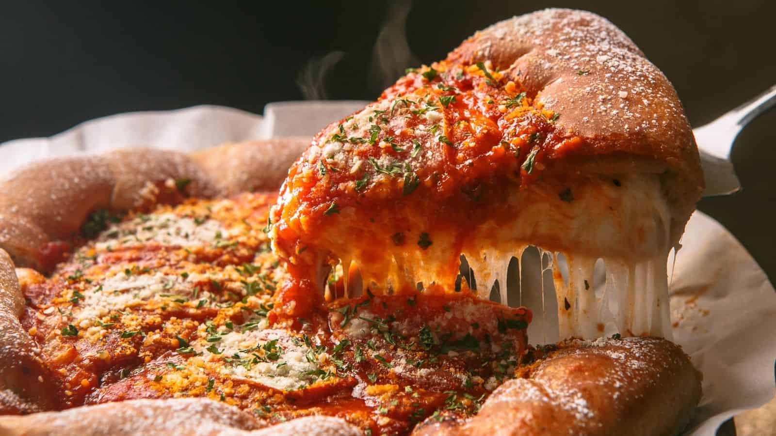 A deep-dish pizza slice is being lifted from the whole pizza, showing melted cheese stretching from the crust. The pizza is topped with tomato sauce, herbs, and grated cheese. Steam is visible, indicating it is hot.