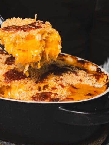 A spoon scoops a portion of baked macaroni and cheese from an oval black dish. The dish is topped with a golden-brown breadcrumb crust. A teal cloth rests beside the dish on a white surface.