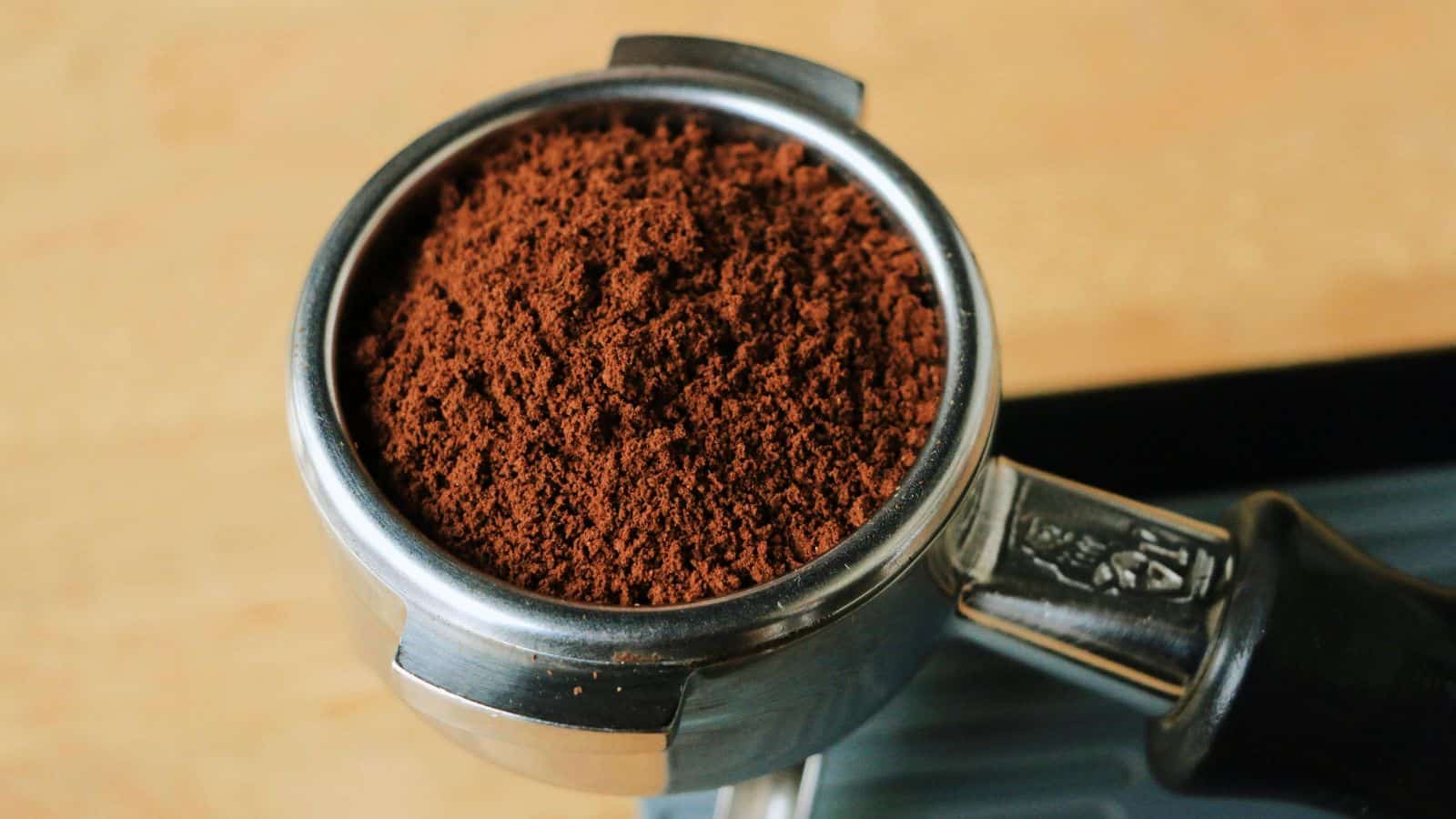 Close-up of a portafilter filled with finely ground coffee, ready for brewing espresso. The background is a light wooden surface.