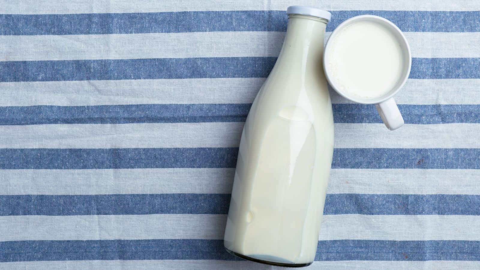 A glass bottle filled with milk lies on a blue and white striped fabric. Beside it, a white cup filled with milk is placed on its side. The setting suggests a simple, rustic presentation.