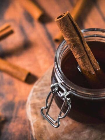 A glass jar filled with dark liquid and a cinnamon stick sits on a wooden surface. Two additional cinnamon sticks are laid nearby, next to a small bowl of ground cinnamon placed on a piece of burlap. The jar's lid is open.
