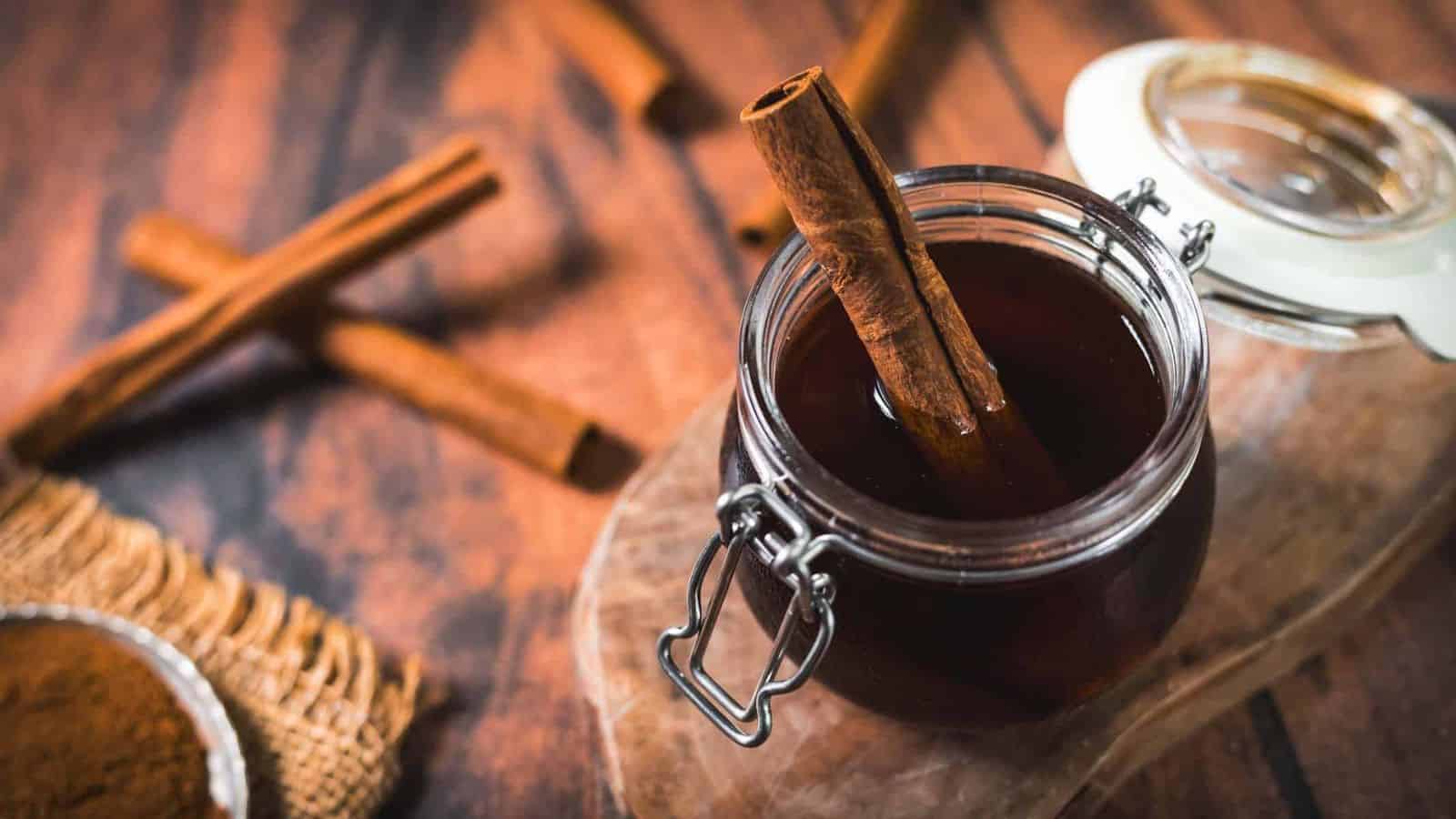 A glass jar filled with dark liquid and a cinnamon stick sits on a wooden surface. Two additional cinnamon sticks are laid nearby, next to a small bowl of ground cinnamon placed on a piece of burlap. The jar's lid is open.