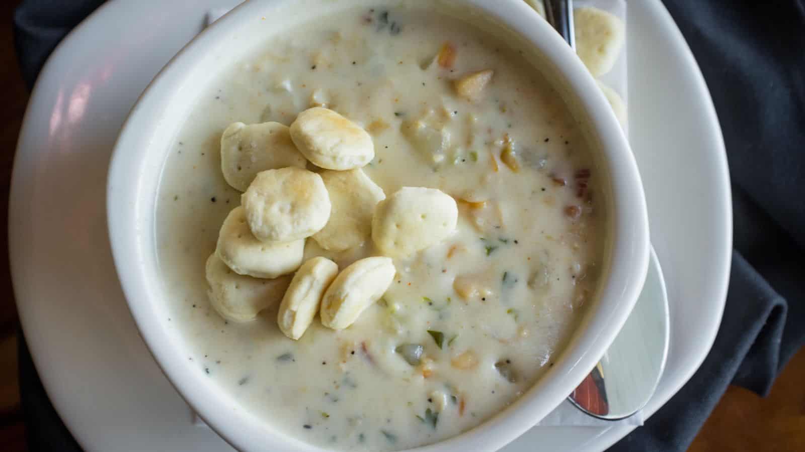 A bowl of creamy clam chowder sits on a white plate, topped with a handful of round oyster crackers. The soup contains visible bits of clams, herbs, and vegetables. A spoon rests on the plate beside the bowl.
