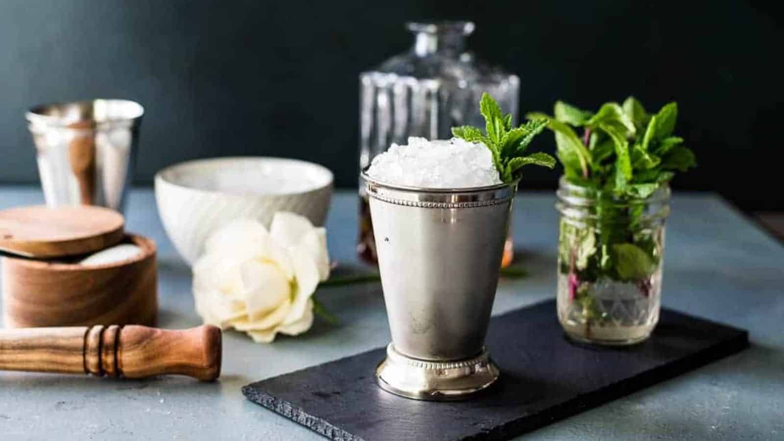 A mint julep in a silver mint julep cup with sprigs of mint and a decanter of bourbon and other ingredients in the background.