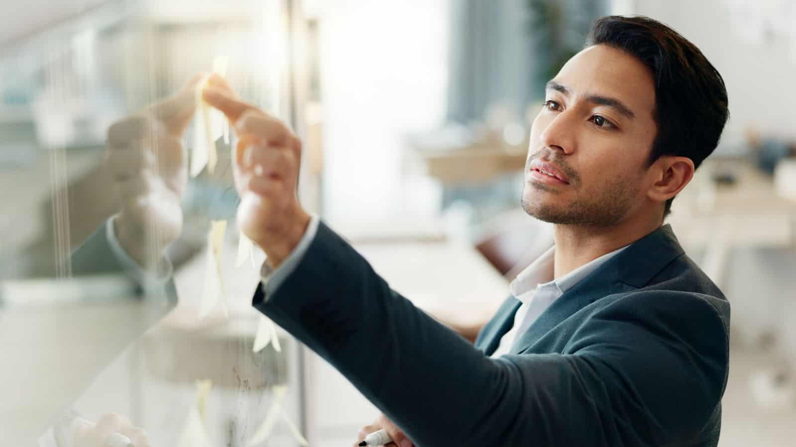 A man in a suit writes on a glass board with sticky notes, focusing intently. The background shows a bright office setting.