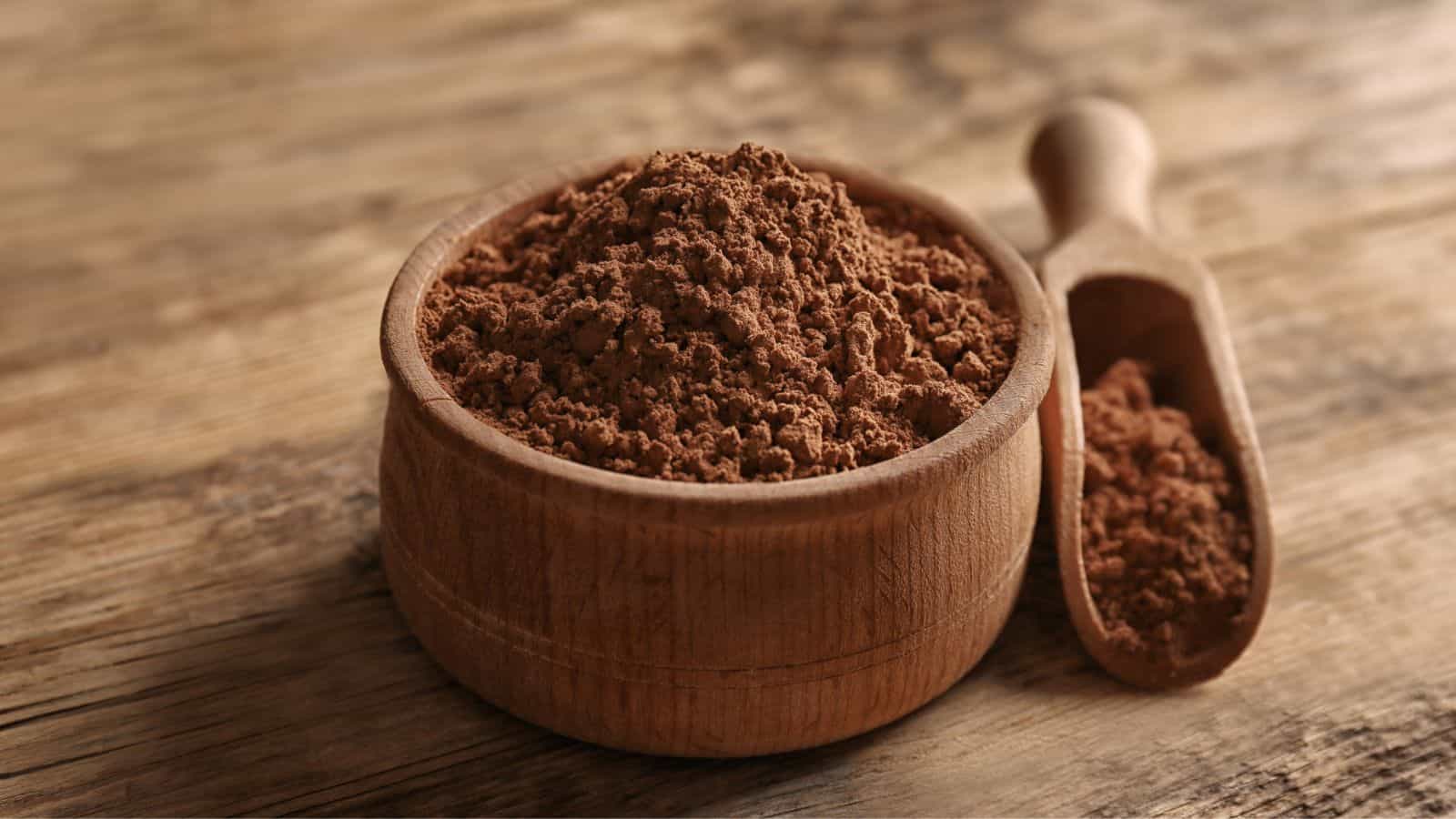 A wooden bowl filled with cocoa powder sits on a textured wooden surface. Next to the bowl is a wooden scoop, also containing some cocoa powder. The scene features warm tones and natural materials.
