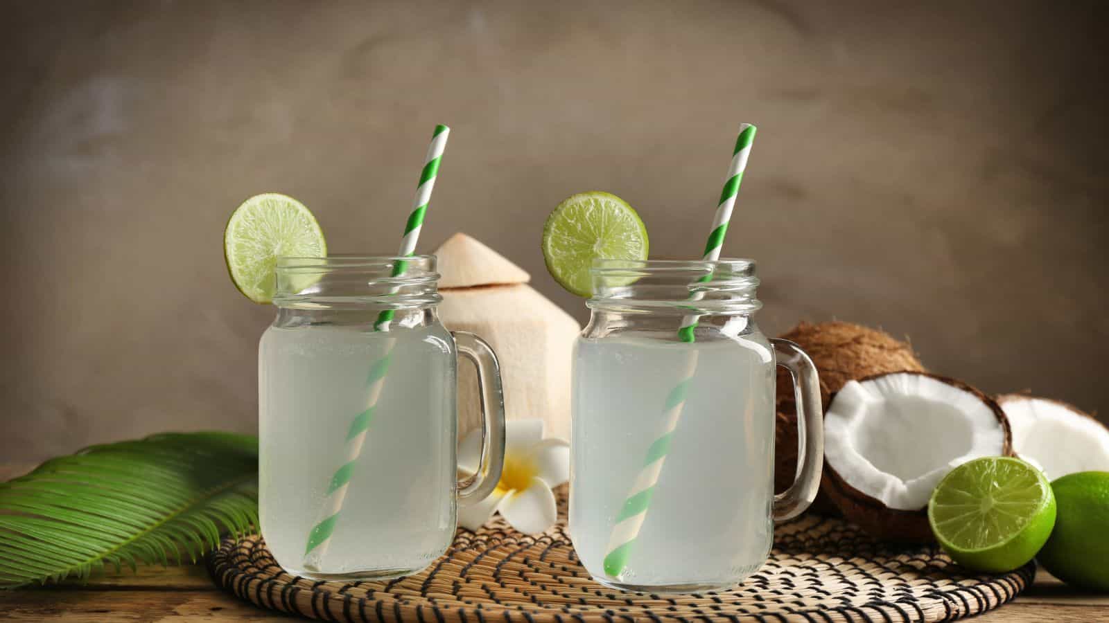 Two mason jar glasses filled with coconut water, each garnished with a lime slice and a green-striped straw. A coconut, lime, and a tropical leaf are in the background, placed on a woven mat.
