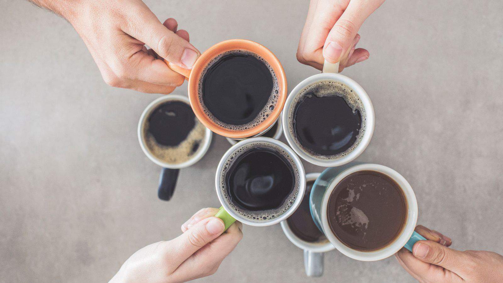 Six hands holding coffee mugs come together in a toast. The mugs contain black coffee and are of various colors, including orange, yellow, green, and black, set against a neutral background.