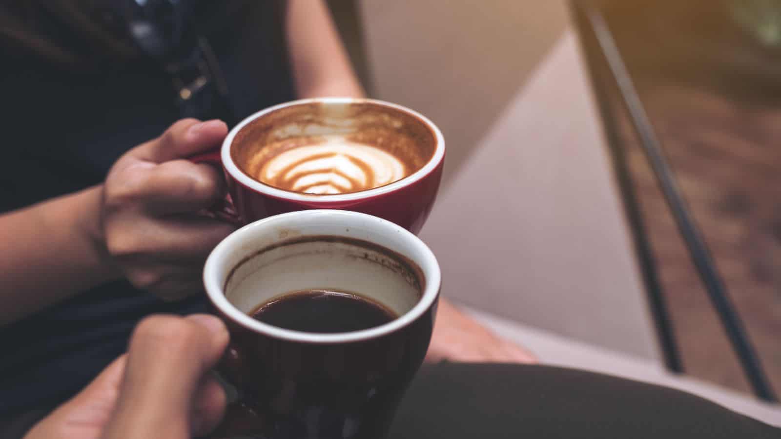 Two people clink ceramic mugs. One mug contains black coffee, and the other has a latte with foam art. They are seated, with sunlight illuminating the scene.