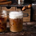 Two glass jars of iced coffee are on a textured surface; one has cream swirling in it, while the other has foam on top. Coffee beans and cinnamon sticks are in the background, alongside a metallic espresso maker.