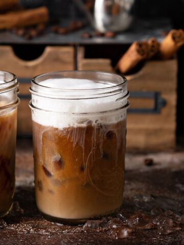Two glass jars of iced coffee are on a textured surface; one has cream swirling in it, while the other has foam on top. Coffee beans and cinnamon sticks are in the background, alongside a metallic espresso maker.