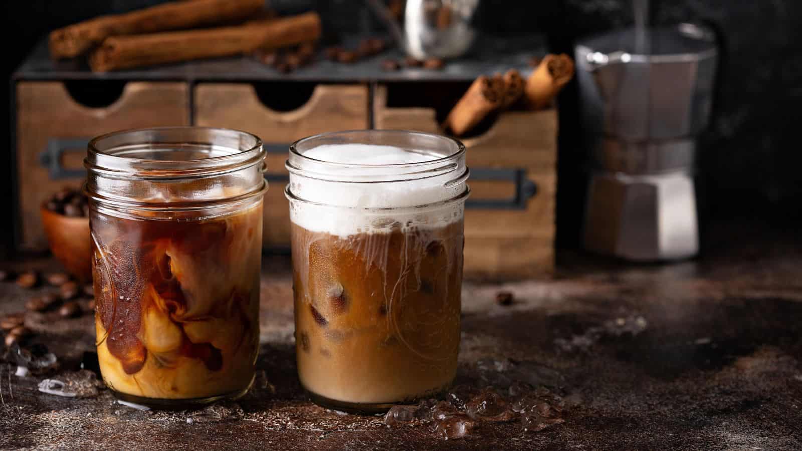 Two glass jars of iced coffee are on a textured surface; one has cream swirling in it, while the other has foam on top. Coffee beans and cinnamon sticks are in the background, alongside a metallic espresso maker.