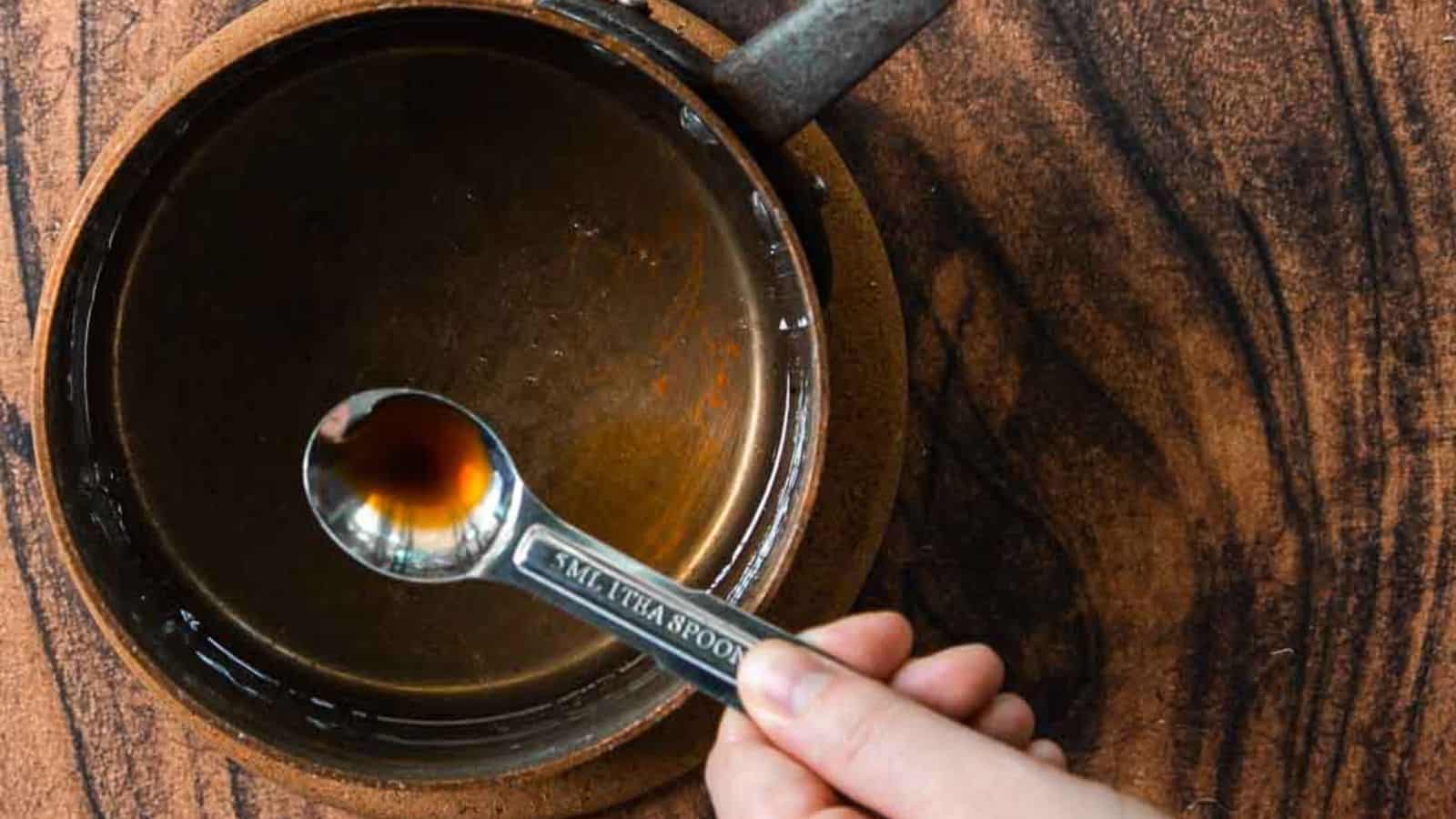 A hand holds a measuring spoon containing dark liquid over a brown ceramic bowl filled with similar liquid. The bowl sits on a wooden surface, and the spoon reads "1 TSP.