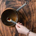 A hand holds a metal measuring spoon filled with a liquid over a brown saucepan. The saucepan is set on a wooden surface with distinct swirling patterns. The person is wearing a colorful braided bracelet on their wrist.