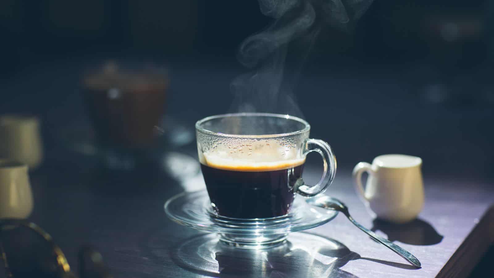 A clear glass cup of steaming coffee sits on a glass saucer with a metal spoon next to it. Soft light highlights the steam rising from the drink. A small white creamer pitcher is visible in the background on a dark surface.