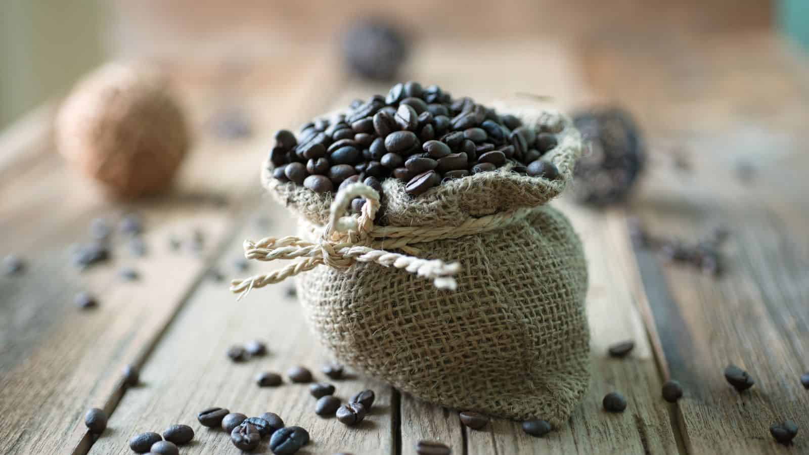 A small burlap sack filled with dark coffee beans sits on a wooden surface. Additional coffee beans are scattered around the sack. The background is softly blurred, featuring more sacks of beans.