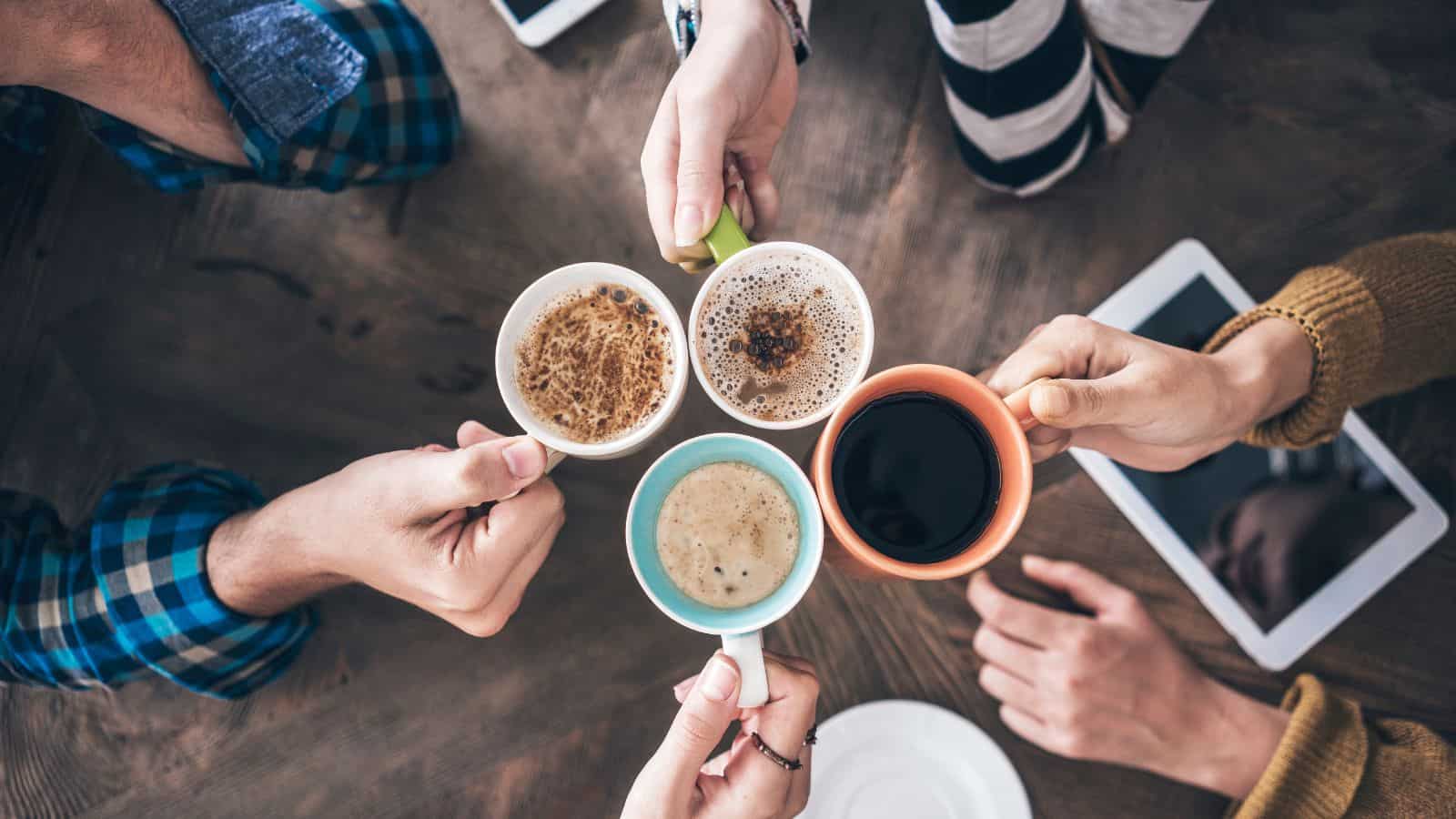 Four people hold out coffee mugs over a wooden table. The mugs contain different beverages, including coffee and frothy drinks. A tablet and a saucer are partially visible on the table. The group wears casual clothing.