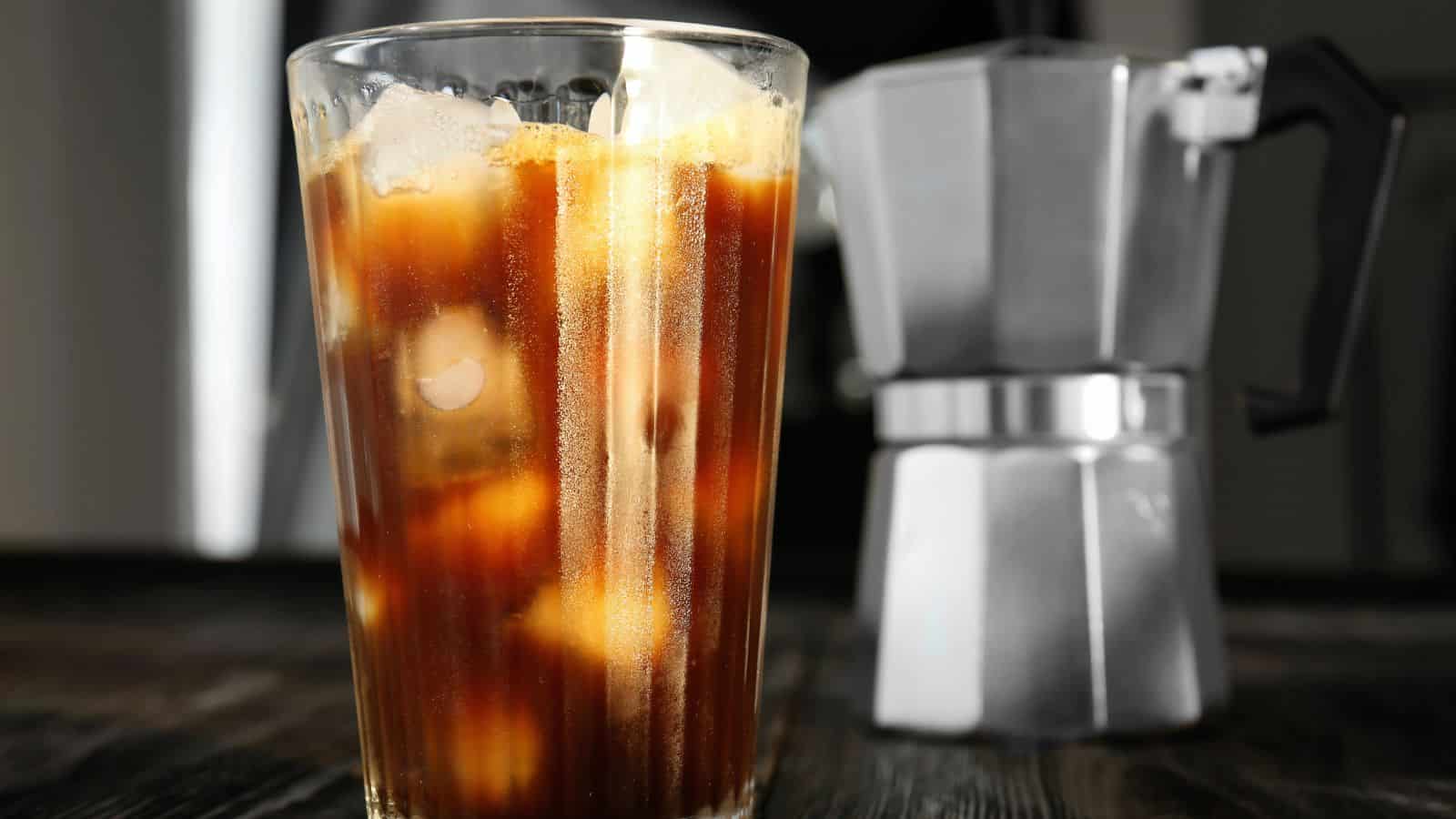 A glass of iced coffee filled with ice cubes is placed on a dark wooden surface. In the blurred background, there is a silver Moka pot. The image captures a refreshing beverage next to a classic coffee maker.