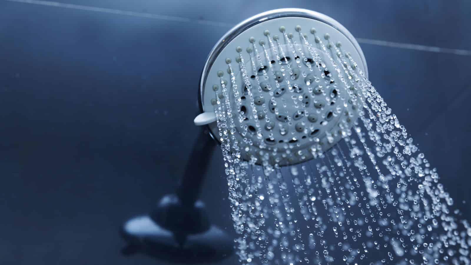 Close-up of a showerhead spraying water. The showerhead is metallic with multiple water jets, releasing streams of water against a dark, blurred background. The water droplets are captured in mid-air.