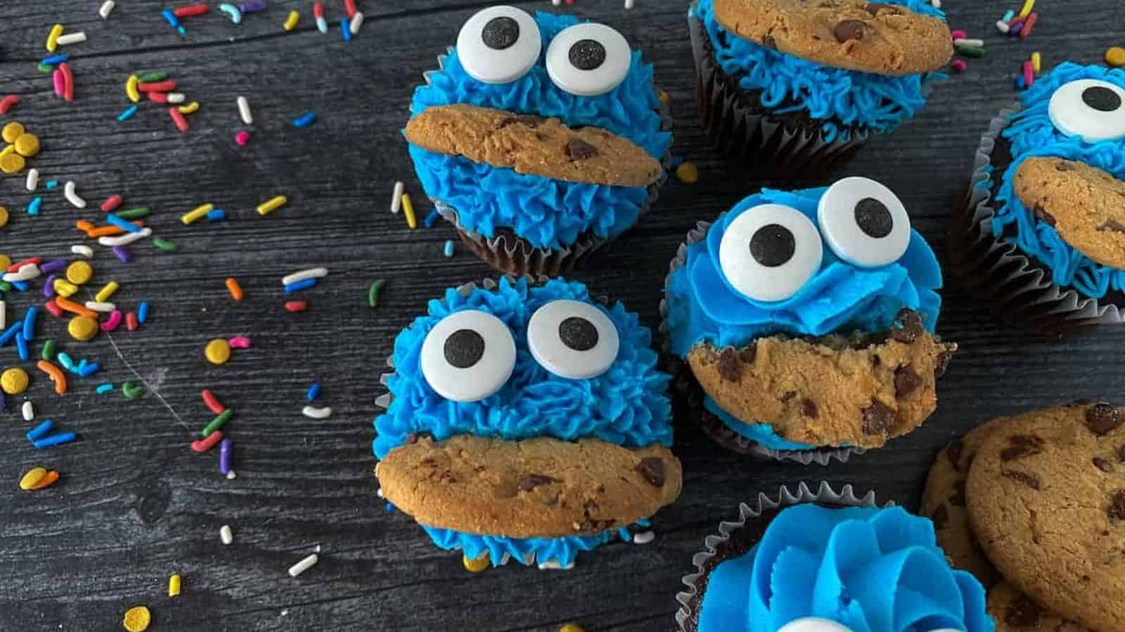Cupcakes decorated to resemble a playful character, featuring blue icing, large googly eyes, and chocolate chip cookies as mouths. Colorful sprinkles are scattered on the dark wooden surface around them.