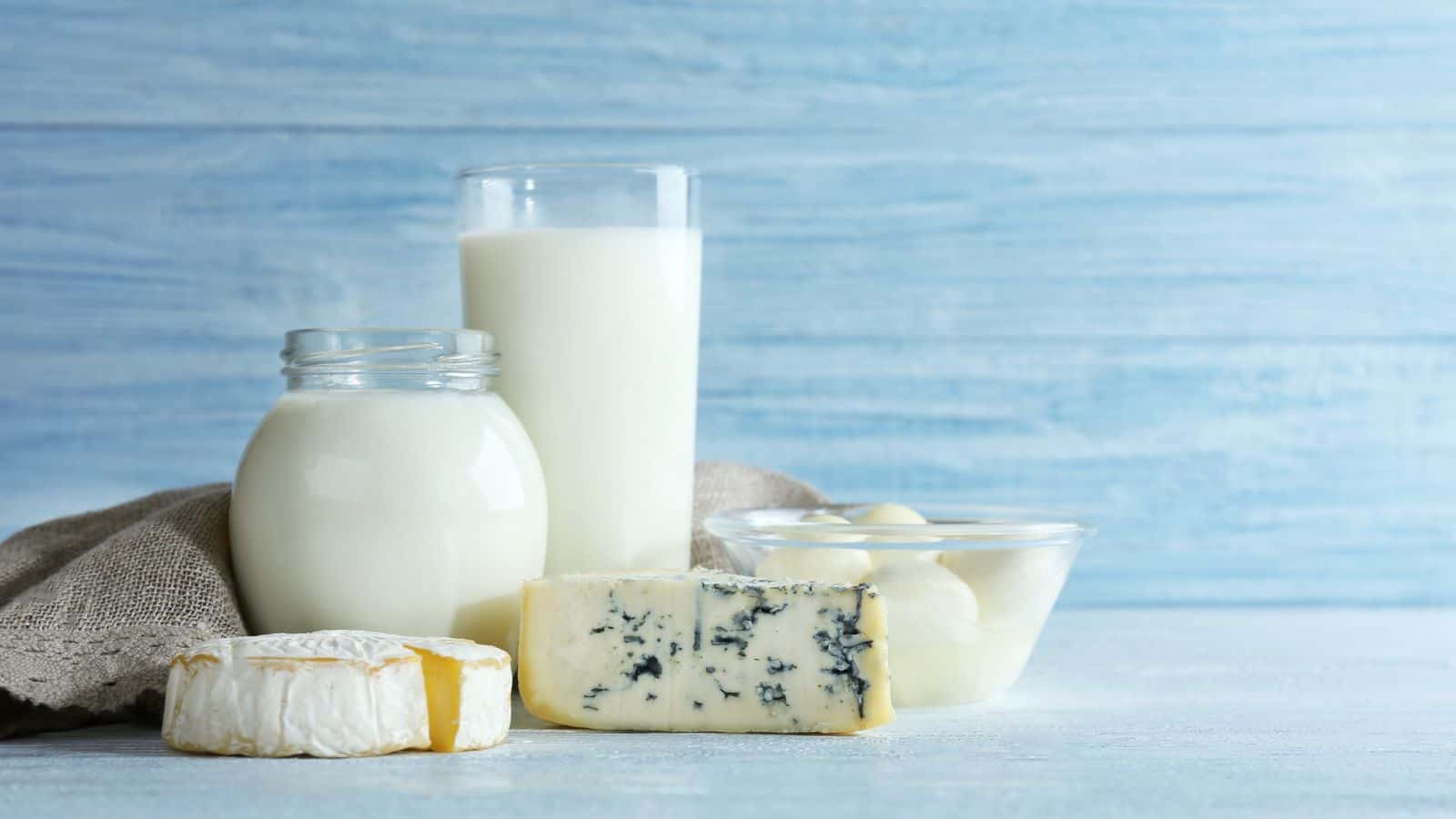 A selection of dairy products is displayed on a light wooden surface. There is a jar and glass of milk, a wheel of brie, a wedge of blue cheese, and a bowl of small mozzarella balls. A beige cloth is partially visible behind the items.