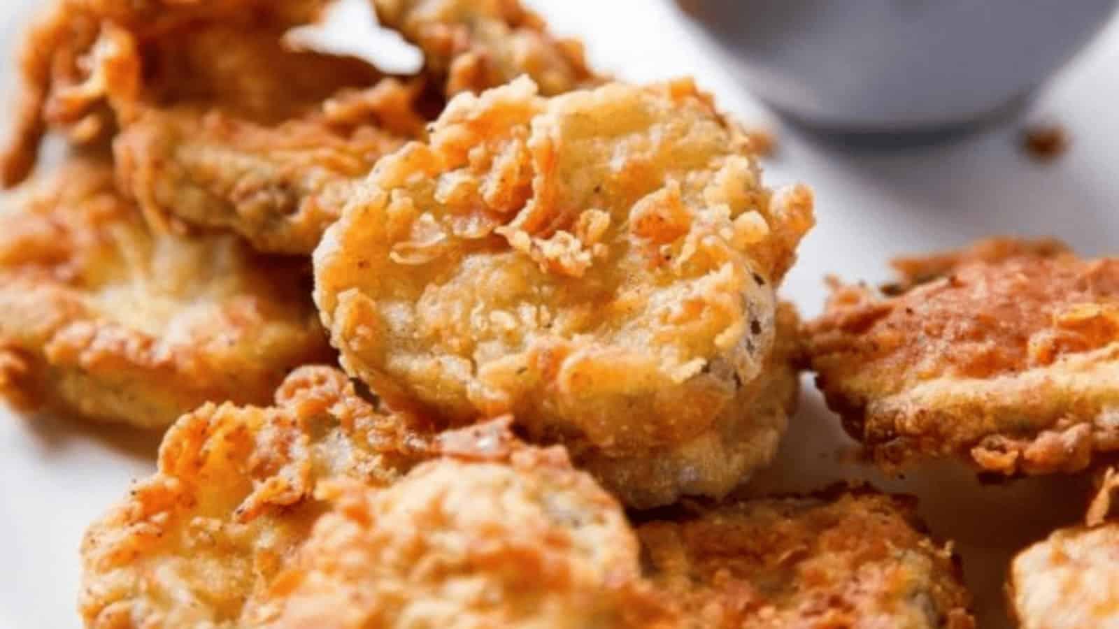 Close-up of golden, crispy fried picklesโan iconic American snackโserved on a white plate with a dipping sauce in the background.