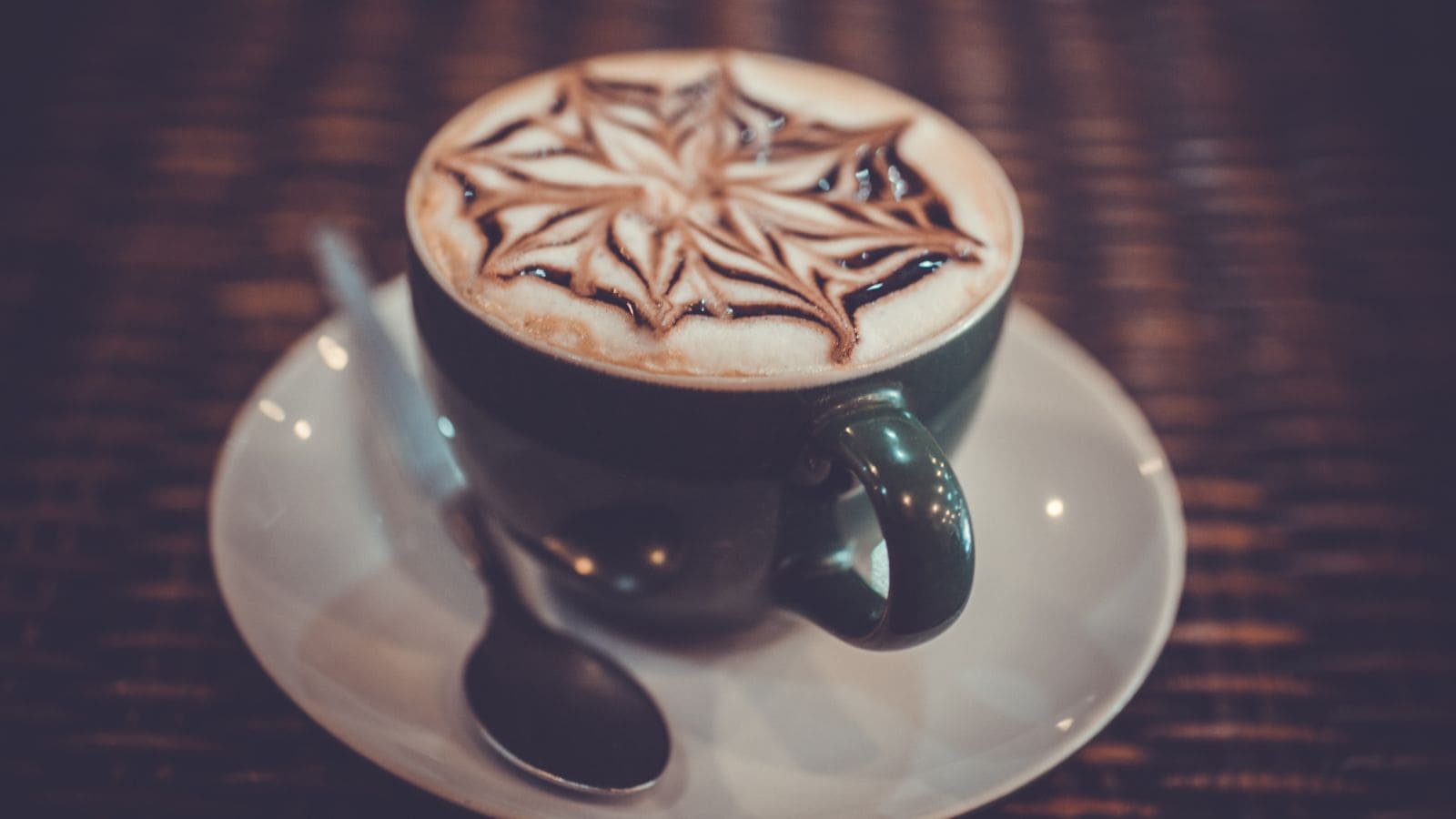 A green cup filled with a frothy coffee topped with an intricate flower pattern in chocolate syrup. It is placed on a white saucer with a silver spoon on a woven surface.