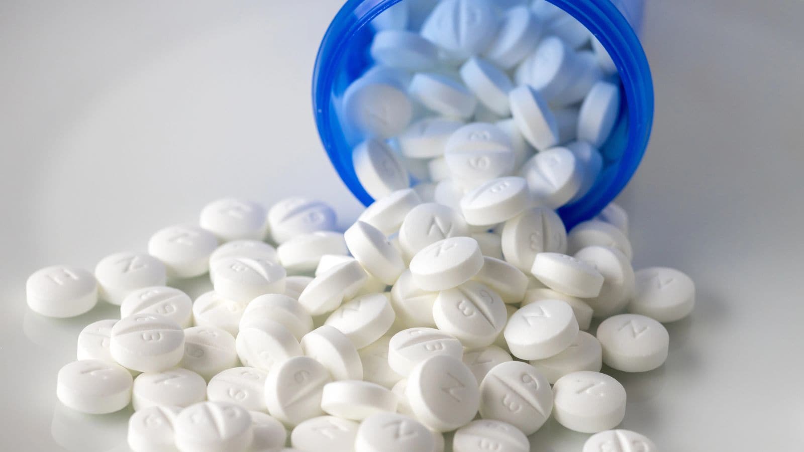 White pills spill from a blue open bottle onto a white surface. The pills are round and have markings, some with the letter 'N'.