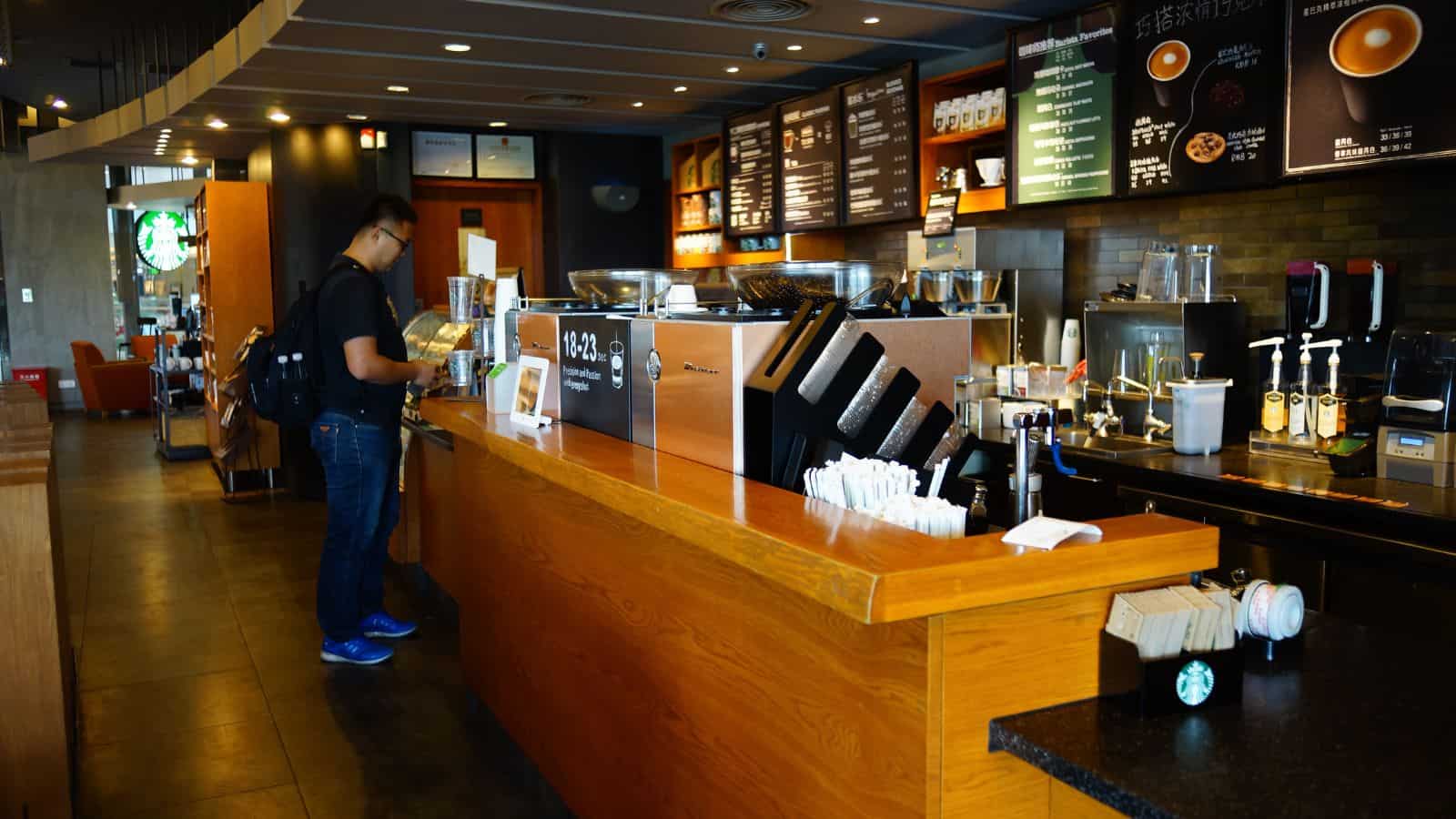 A person stands at the counter in a coffee shop, looking at the menu. The counter features various coffee-making equipment and supplies. The interior has wooden accents and overhead lighting. Wall menus display drink options with images and text.