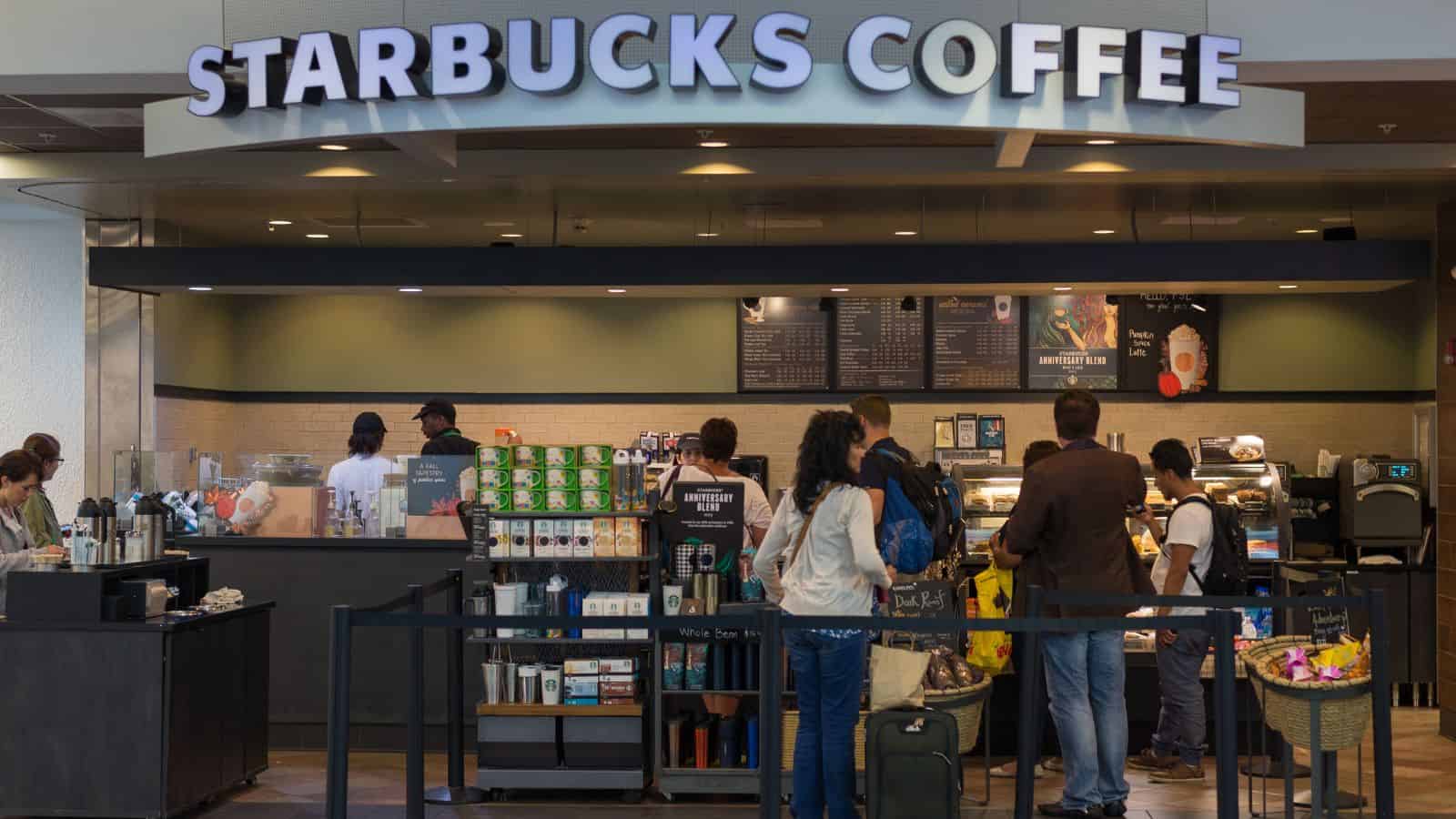Customers wait in line at a Starbucks coffee shop. The counter displays various products, and the menu is visible above. People are ordering or preparing drinks in the background. The store is well-lit and has a modern design.