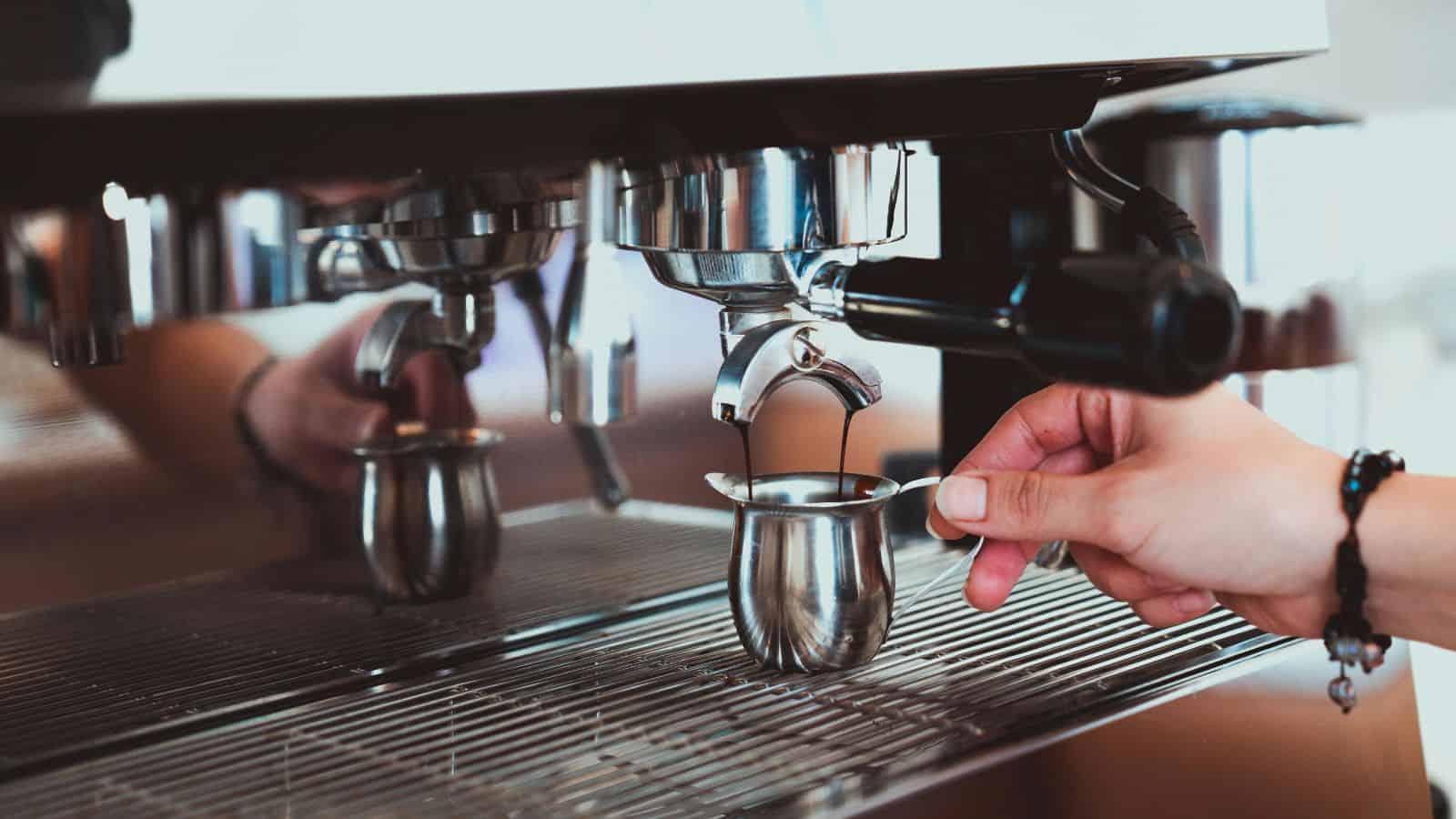 A hand operates an espresso machine, with coffee being brewed into a small metal pitcher. The machine features reflective surfaces and several group heads.