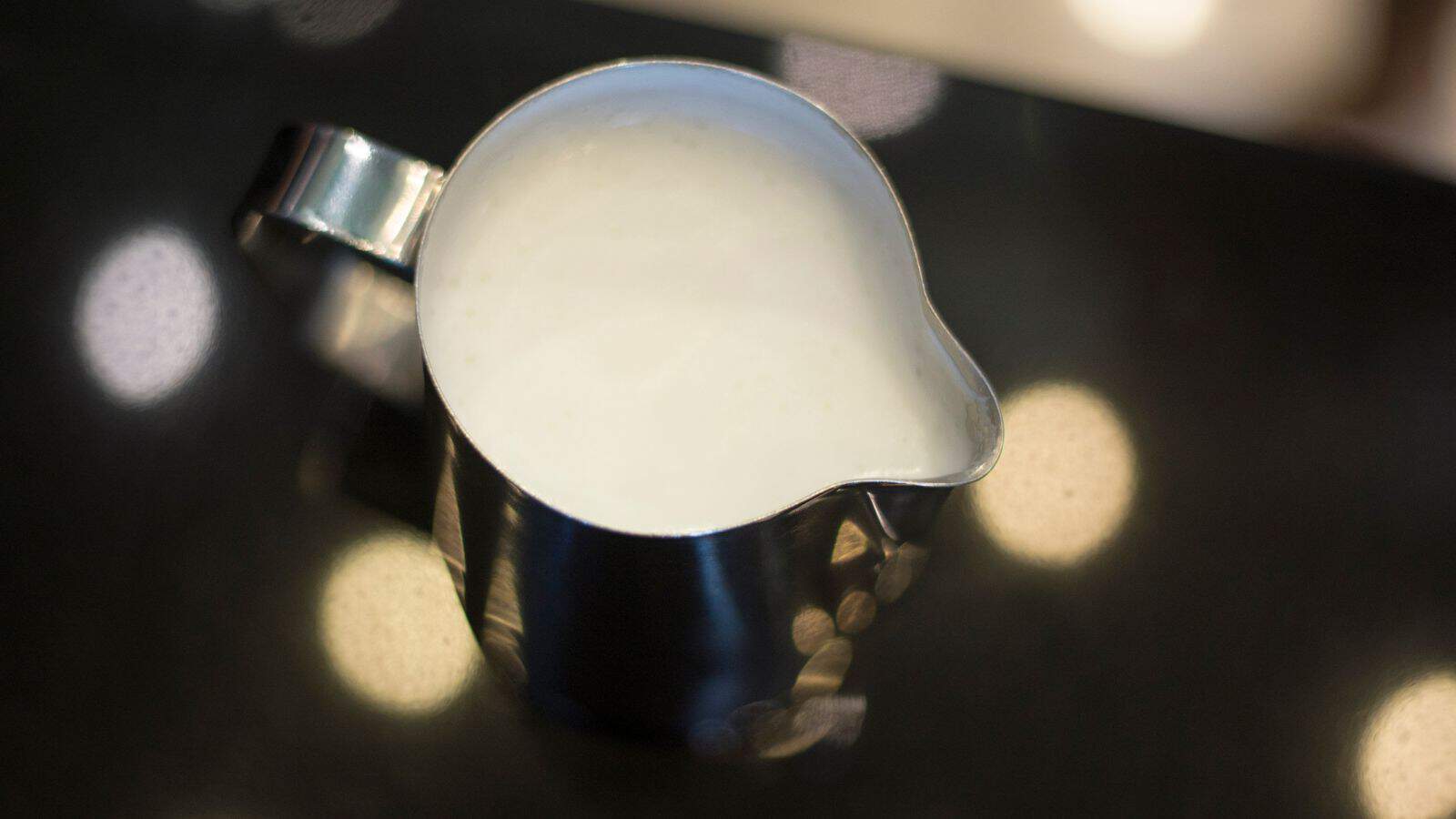 A metal pitcher filled with steamed milk sits on a dark countertop. The milk has a frothy texture, and the handle of the pitcher is on the left side. Bright circular reflections are visible on the surface around the pitcher.