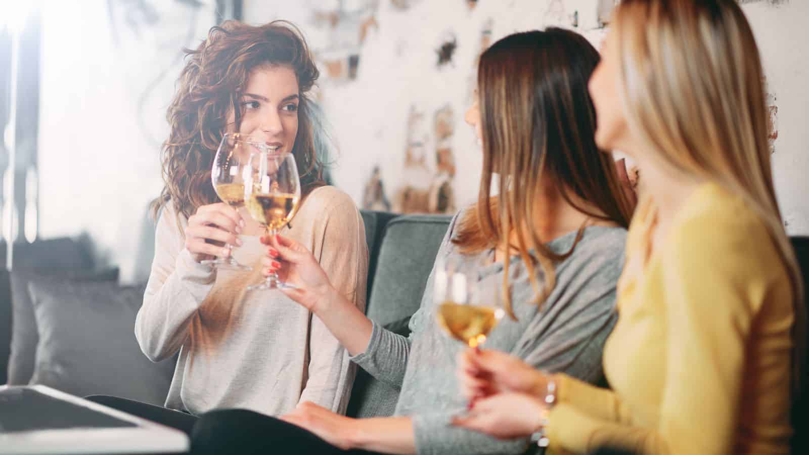 Three women sit together on a couch, holding wine glasses and smiling. They appear to be engaged in conversation in a relaxed, social setting. The background features a light-colored wall with some textured details.