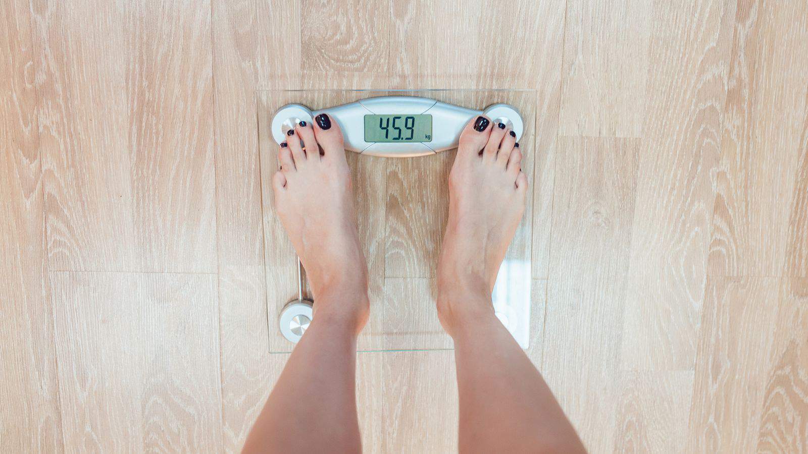 A person stands on a digital scale displaying a weight of 45.9 kilograms. The scale is placed on a light wooden floor, and the person's toes are visible, painted with dark nail polish.