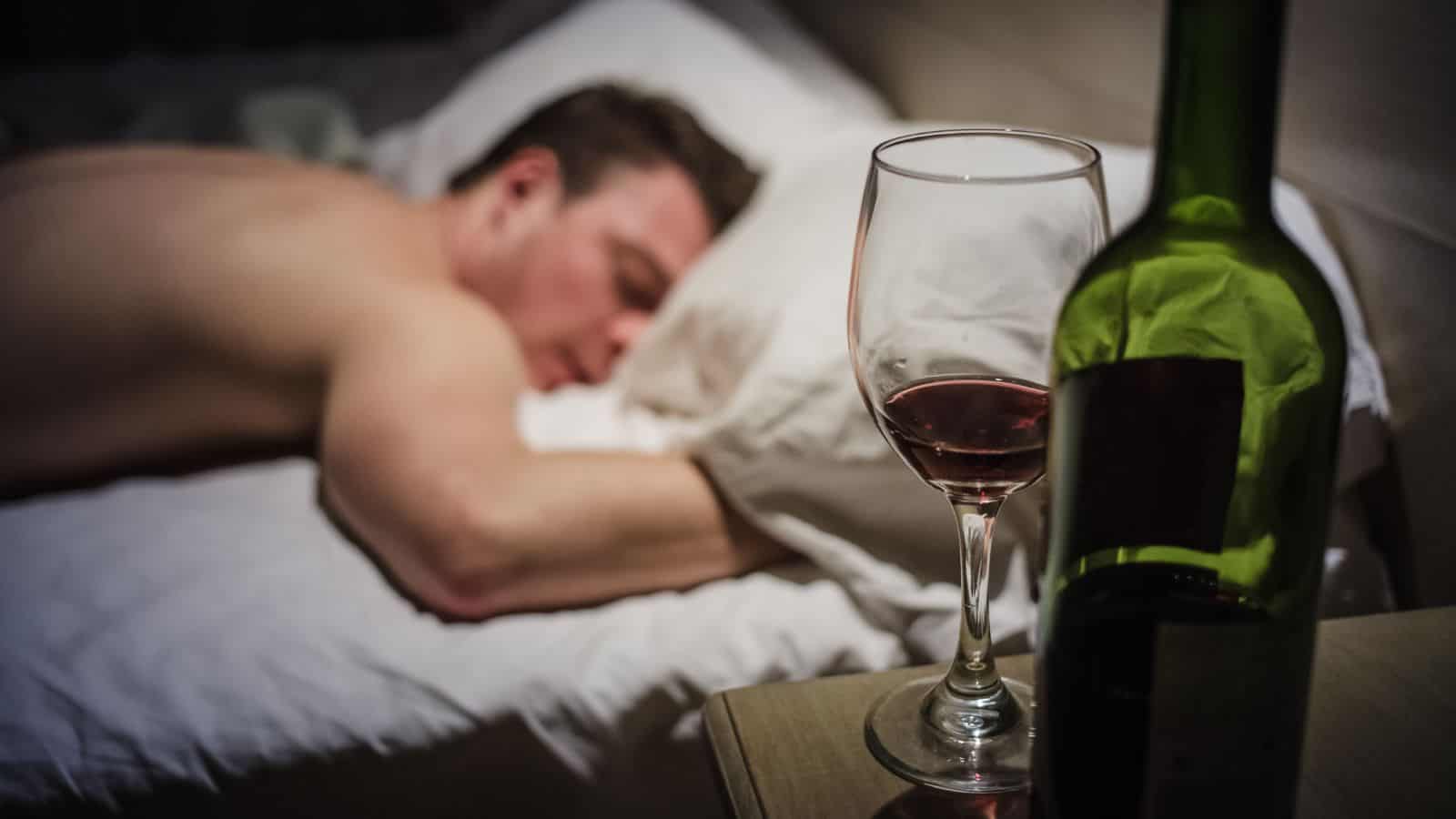 A shirtless man is sleeping on a bed with his head on a white pillow. In the foreground, there is a glass of red wine and a green wine bottle on a wooden bedside table. The room is dimly lit.