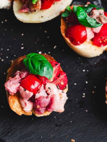 Slices of baguette topped with diced tomatoes, bacon, and basil leaves are arranged on a black slate surface. The ingredients are seasoned with cracked black pepper.