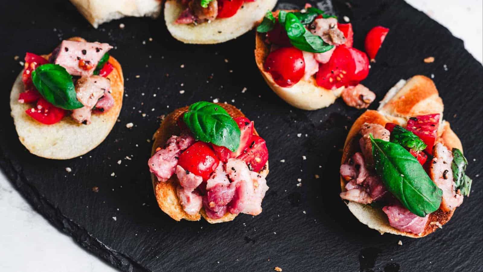 Slices of baguette topped with diced tomatoes, bacon, and basil leaves are arranged on a black slate surface. The ingredients are seasoned with cracked black pepper.