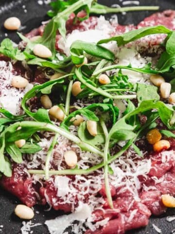 A black plate features beef carpaccio topped with arugula, grated Parmesan cheese, and pine nuts. The thinly sliced meat is arranged with the garnishes spread evenly across its surface.
