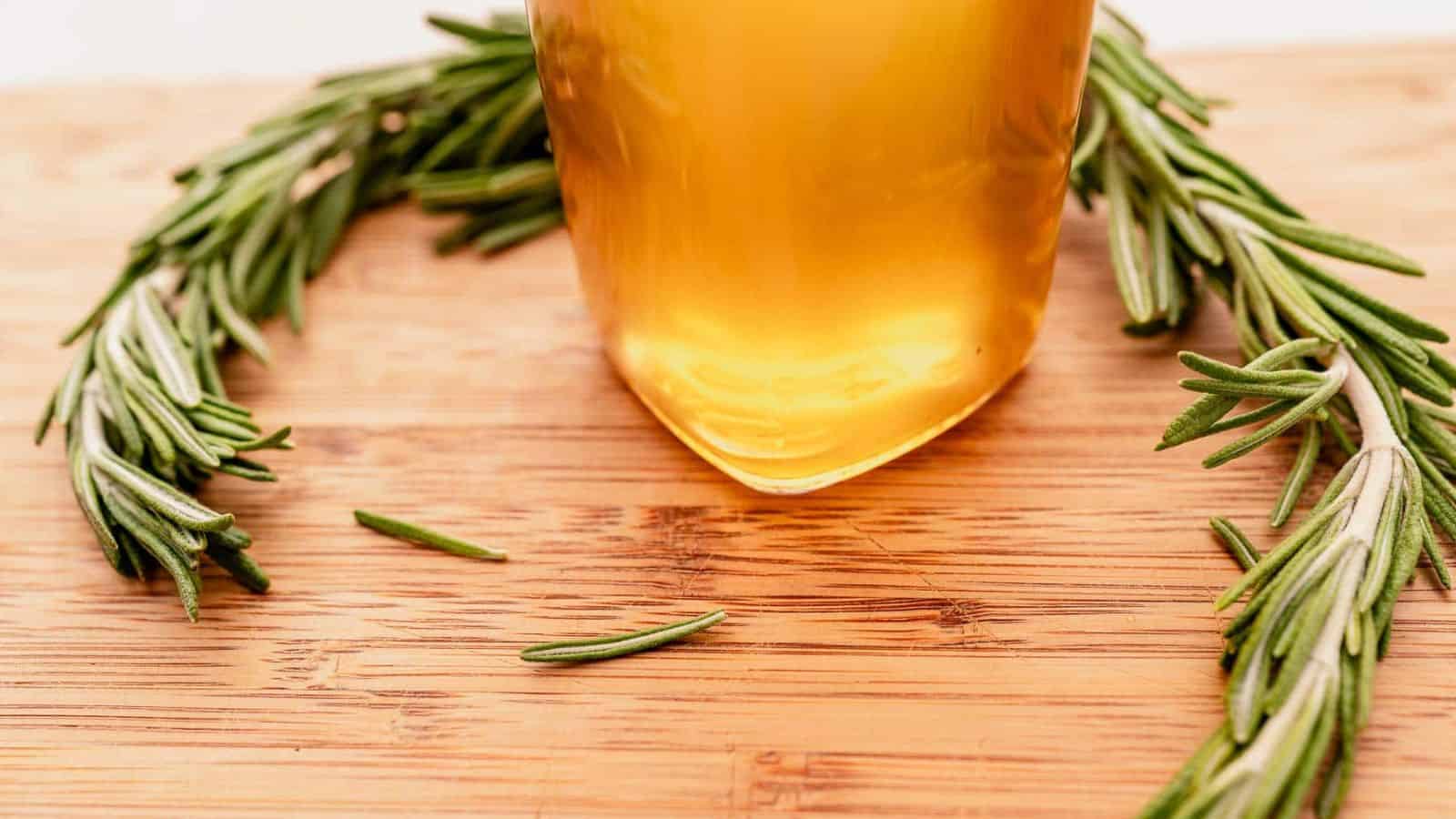 A glass of amber-colored liquid on a wooden surface, surrounded by a sprig of fresh rosemary.