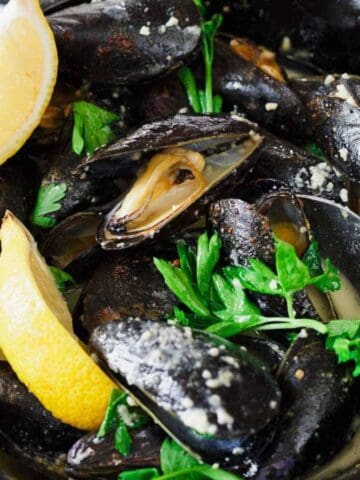 A skillet contains cooked mussels in a light sauce, garnished with parsley. A lemon wedge is placed inside, and a spoon rests in the dish. Partially visible bread rolls are on the side.