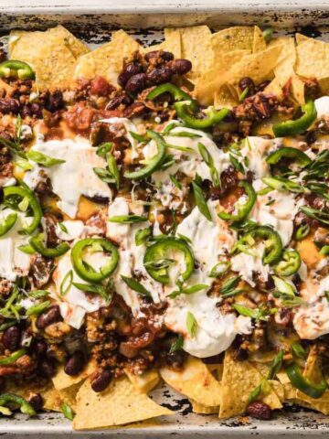 A sheet pan filled with nachos topped with melted cheese, ground meat, kidney beans, jalapeño slices, chopped green onions, and dollops of sour cream. The nachos are placed on a marble surface.