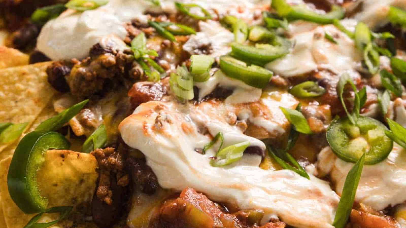 A close-up of a hand holding a tortilla chip topped with melted cheese, meat, beans, and green onions. The chip is pulled from a tray of loaded nachos, with strings of cheese stretching from the tray.