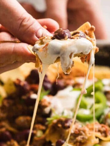 A close-up of a hand holding a tortilla chip topped with melted cheese, meat, beans, and green onions. The chip is pulled from a tray of loaded nachos, with strings of cheese stretching from the tray.